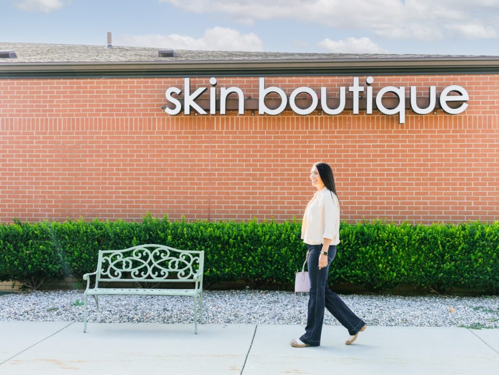 A smiling brunette woman walks on the sidewalk under the Skin Boutique sign. She's coming to our med spa for microneedling in Oakland County, MI.